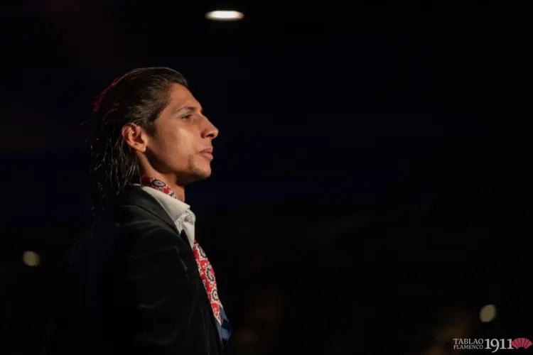 Plano medio-corto de perfil del bailaor flamenco Ricardo Fernández "El Tete", capturado con una expresión introspectiva y concentrada en el escenario oscuro de Tablao Flamenco 1911. Viste una chaqueta de terciopelo negro sobre una camisa blanca de cuello abierto, complementada con un pañuelo de colores estampado. La iluminación dramática del escenario resalta su perfil. El fondo es completamente oscuro. El logotipo de Tablao Flamenco 1911 es visible en la esquina inferior derecha.