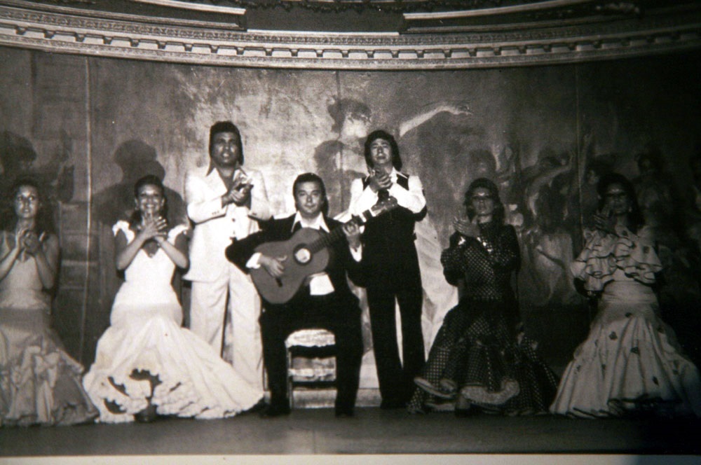 Fotografía vintage en blanco y negro del tocaor Antonio Arenas tocando la guitarra flamenca en el centro del escenario del antiguo Tablao Villa Rosa en los años 60. Está sentado y rodeado por un cuadro flamenco de cantaores y bailaoras vestidos de época que le acompañan con las palmas.