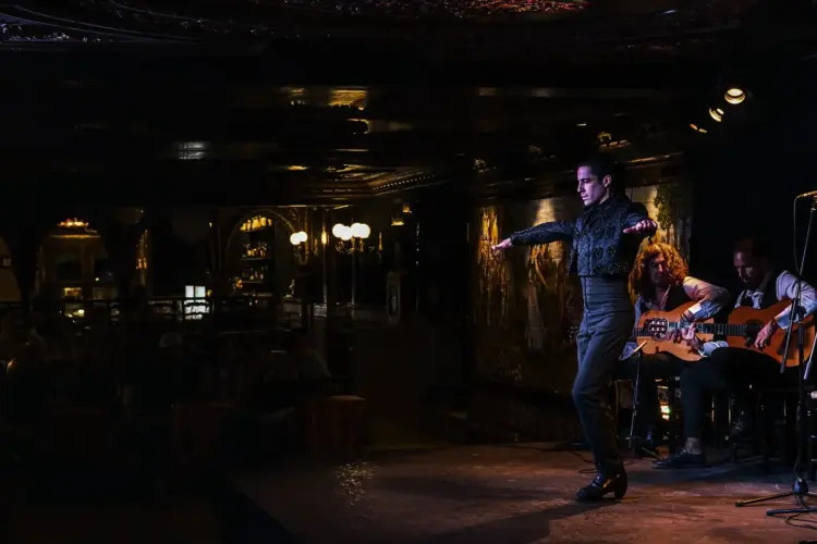 Un bailaor flamenco en el centro de un escenario de madera noble, en plena actuación con fuerza y pasión. Detrás de él, dos guitarristas tocan sentados. El ambiente es oscuro e íntimo, con iluminación de escenario cálida, típico de un tablao flamenco tradicional en Madrid.