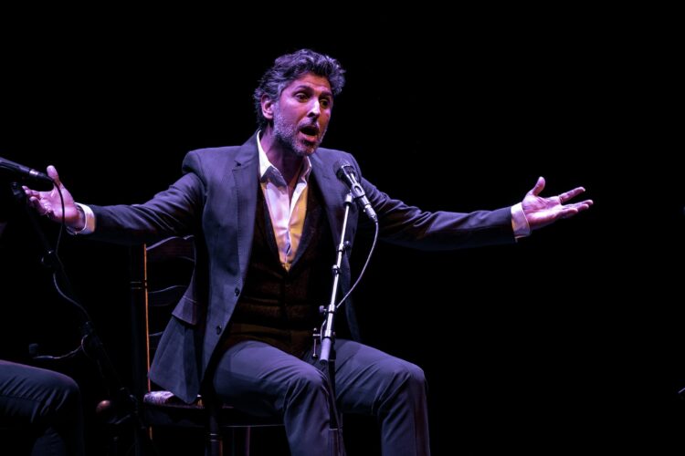 El cantaor flamenco Arcángel interpretando un fandango con profunda emoción y los brazos abiertos sobre un escenario, acompañado por dos guitarristas flamencos a sus lados bajo una iluminación teatral dramática.